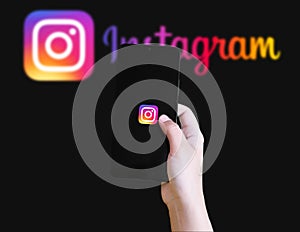 Hand holding smartphone with Instagram app loading screen in front of blurred Instagram logo illustrative