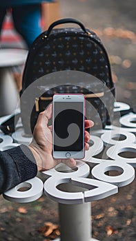 Hand holding smartphone on unique table, blurred background, stylish backpack adds character.