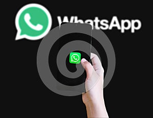 Hand holding smartphone with WhatsApp app icon on screen and logo in background illustrative editorial