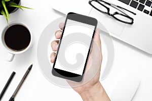 Hand holding smartphone with white blank screen over white office desk table with supplies