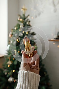 Hand holding stylish christmas vintage ornament on background of christmas tree in lights in festive boho room. Decorating