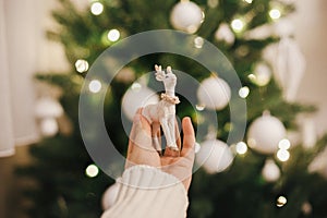 Hand holding stylish reindeer toy on background of christmas tree lights. Beautiful christmas deer toy in hand in festive