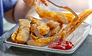 Hand holding tasty baked potato wedges with fork and dipping in ketchup in a plate at restaurant