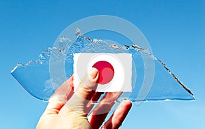 A hand holding thin transparent piece of ice in the shape of an iceberg with the flag of the Japan against a blue sky. The concept