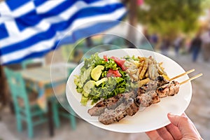 Hand holding a traditional greek pork souvlaki plate with fried potatoes and green salad.