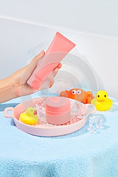 A hand holding a tube of pink cosmetics, a jar of lotion, silicone bath toys, a bath sponge, ceramic tray and ice are placed on a