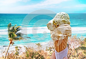 Hand is holding the vanilla ice cream. In the background is blue sky, sea nd sandy beach. There are palms