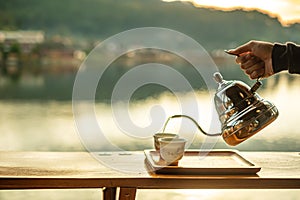 Hand holding vintage teapot and pouring hot tea to cup on wood table against lake view background at coffee shop in the morning