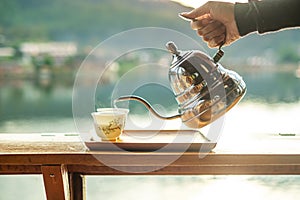 Hand holding vintage teapot and pouring hot tea to cup on wood table against lake view background at coffee shop in the morning