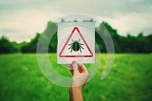 Hand holding a warning sign for ticks parasite danger over the park green lawn background. Different bug bites, health risk,