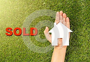 Hand holding white house and word Sold on green grass in conceptual image of property business