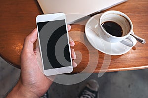 Hand holding white mobile phone with blank black desktop screen with coffee cup and laptop on wooden table in cafe