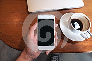 Hand holding white mobile phone with blank black desktop screen with coffee cup and laptop on wooden table in cafe