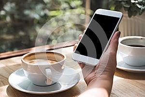 Hand holding white mobile phone with blank black desktop screen with coffee cup on wooden table in cafe