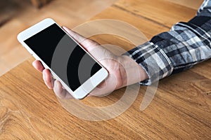 Hand holding white mobile phone with blank black desktop screen on wooden table in cafe