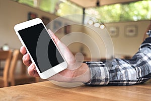 Hand holding white mobile phone with blank black desktop screen on wooden table in cafe