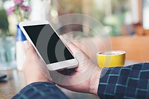 A hand holding white mobile phone with blank black desktop screen and yellow coffee cup on wooden table in modern cafe