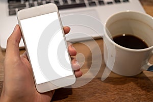 Hand holding white mobile phone with blank desktop screen with coffee cup and laptop on wooden table in cafe