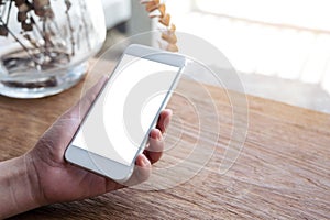 Hand holding white mobile phone with blank desktop screen on wooden table in cafe