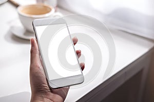 A hand holding white mobile phone with blank screen , coffee cup on white vintage wooden table and curtain background