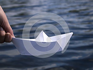 A hand is holding a white paper boat above the water.