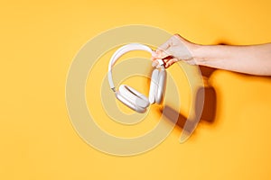 Hand holding white wireless headphones against a yellow background under hard light