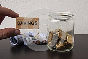 Hand holding wooden block with text Savings. Bank notes and gold coin in a jar background.