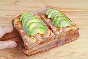 Hand holding a wooden breadboard of grilled cheese toast with tomato and sliced fresh ripe avocado