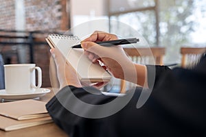 A hand holding and writing on blank notebook with coffee cup on table in cafe