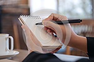 A hand holding and writing on blank notebook with coffee cup on table in cafe