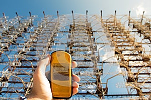 Hand holding a yellow dosimeter indicating radiation on the screen. Enormous Duga radar complex in the background. Warm summer day