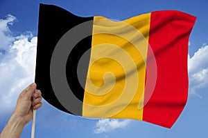 Hand holds against the background of the sky with clouds the colored flag of Belgium on the texture of the fabric, silk with waves