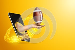 Hand holds American football ball via smartphone on yellow background. Concept for online games, sports broadcasts, sports betting photo