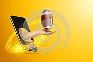 Hand holds American football ball via smartphone on yellow background. Concept for online games, sports broadcasts, sports betting