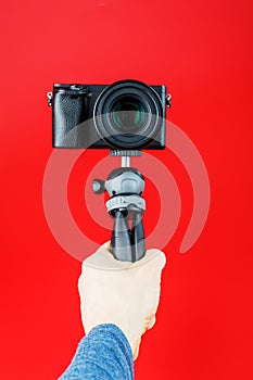 A hand holds a black SLR camera on a tripod, isolated on a red background