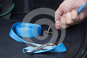 The hand holds a blue ratchet tie in a car trunk.