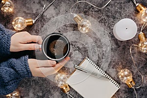 Hand holds cup of coffee on gray table with notebook, pen and candle decorated with led lights.