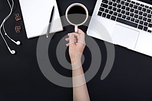 hand holds a cup of coffee on a working black table next to a laptop