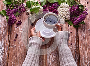 Hand holds a cup of morning coffee with spring lilac flowers branches blossoming on wooden background view from above. Flat lay