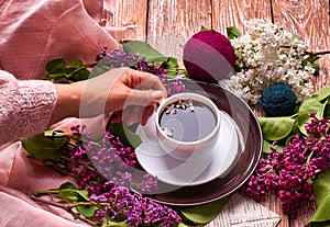 Hand holds a cup of morning coffee with spring lilac flowers branches blossoming on wooden background view from above. Flat lay