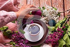 Hand holds a cup of morning coffee with spring lilac flowers branches blossoming on wooden background view from above. Flat lay