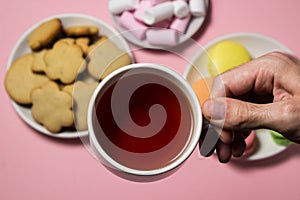The hand holds a cup of tea on the background of sweets. Cup of tea on a pink background.