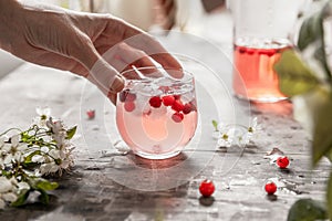 Hand holds a glass of cranberry lemonade with ice and berries on a gray shabby table with flowers and leaves