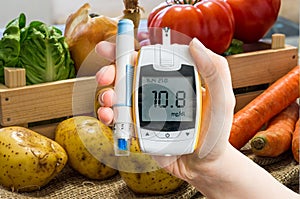 Hand holds glucometer for measuring glucose level. Vegetables in background.