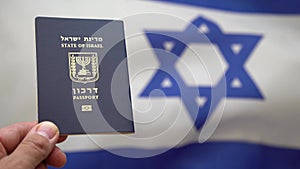 Hand holds an Israeli international passport against the Israeli flag waving in the wind