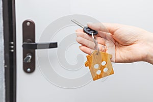 Hand holds the key to the apartment with a wooden house keychain in the background of the entrance door to the flat. The