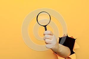 Hand holds a magnifying glass through a torn hole in a paper yellow wall. Search information concept