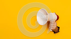 Hand holds a megaphone from a hole in the wall on a yellow background with copy space. Concept of hiring, advertising