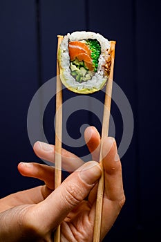 hand holds one piece of Philadelphia sushi roll with smoked salmon, avocado, cucumber and green calve with bamboo