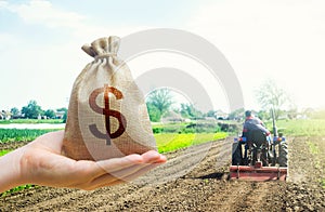A hand holds out a dollar money bag on a background of a field cultivating tractor. Lending farmers for purchase land and seed photo